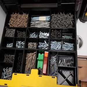 May include: A black plastic toolbox with multiple compartments filled with various types of screws and nails. The toolbox has a yellow handle and a yellow trim around the edges.