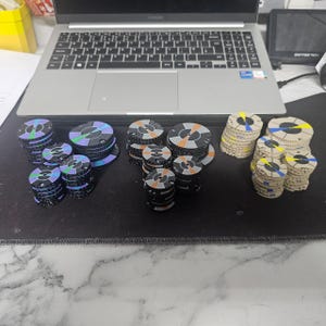 May include: A collection of circular game pieces in various colours, including black, purple, orange, and yellow. The pieces are stacked on a black surface, with a laptop visible in the background. The game pieces appear to be made of cardboard or a similar material.