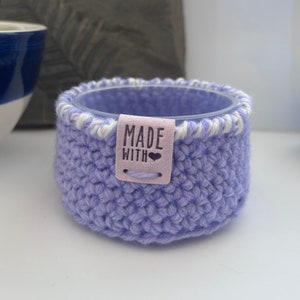May include: A lavender crocheted pot with a white trim and a label that says "MADE WITH" and a heart.