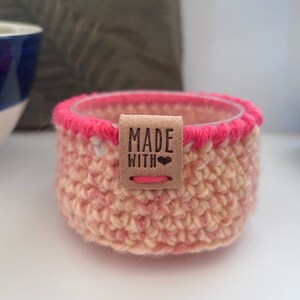 May include: A crocheted pink and cream coloured basket with a brown leather tag that says "MADE WITH" and a heart.