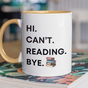 Pode incluir: Caneca de cerâmica branca com alça amarela. A caneca tem texto preto que diz "HI. CAN'T. READING. BYE." e uma pequena ilustração de uma pilha de livros.