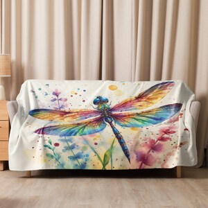 May include: A white throw blanket featuring a watercolour painting of a colourful dragonfly on a white background with green and pink flowers.