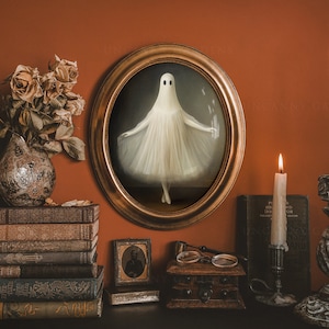 May include: A white ghost figure in a white dress is framed in a gold oval frame. The frame is on a wall with a stack of books, a small wooden box, and two candlesticks with lit candles.