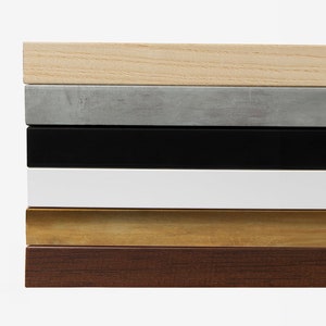 May include: A close-up of six different wood frame samples. The frames are stacked on top of each other, starting with a light wood frame, followed by a gray frame, a black frame, a white frame, a gold frame, and a dark wood frame.