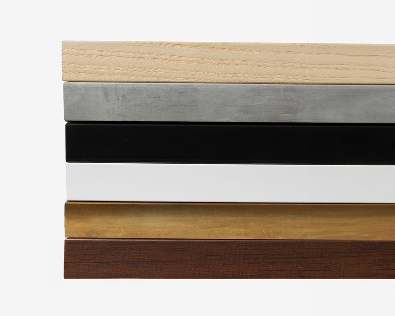 May include: A close-up of six different wood frame samples. The frames are stacked on top of each other, starting with a light wood frame, followed by a gray frame, a black frame, a white frame, a gold frame, and a dark wood frame.