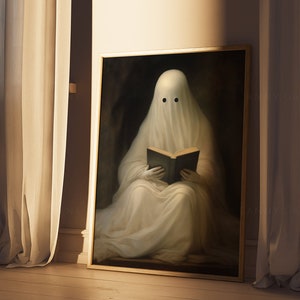 May include: A framed print depicts a ghost reading a book. The ghost is draped in a white sheet with two black eye holes. The artwork has a dark, moody background and is framed in gold. The image is set against a backdrop of beige curtains and a wooden floor.