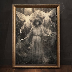 May include: A framed black and white photograph of a woman in a white dress standing in a forest. She is surrounded by a veil of fabric and two large butterfly wings. The image has a vintage feel.