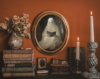 May include: An oval framed portrait of a ghost reading a book, with two black eyes. The image is surrounded by lit candles, antique books, a vase of dried flowers, and a small framed photograph. The scene is set against a burnt orange wall.