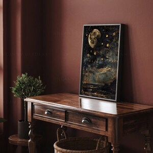 May include: A framed print of a moon and stars against a dark blue background. The moon is a pale yellow color and the stars are gold. The print is hanging on a dark red wall above a wooden table.