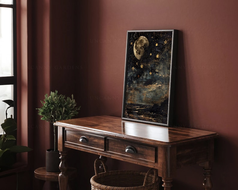 May include: A framed print of a moon and stars against a dark blue background. The moon is a pale yellow color and the stars are gold. The print is hanging on a dark red wall above a wooden table.