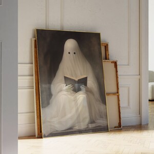 May include: A framed artwork depicts a ghost reading a book. The ghost is draped in a white sheet with two black eye holes. The artwork has a gold frame and is leaning against a wall. The background is a dark, moody color.