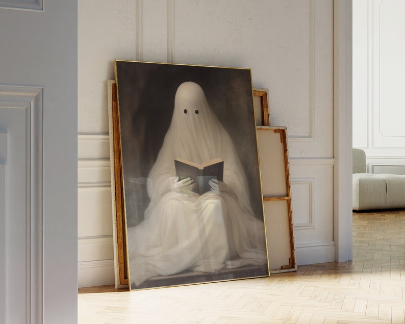 May include: A framed artwork depicts a ghost reading a book. The ghost is draped in a white sheet with two black eye holes. The artwork has a gold frame and is leaning against a wall. The background is a dark, moody color.