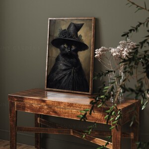 May include: A black cat wearing a witch's hat with black roses and a black cape, framed in a dark wood frame, sitting on a wooden table with a vase of white flowers.