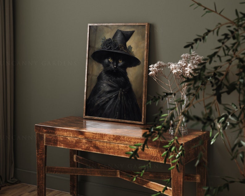 May include: A black cat wearing a witch's hat with black roses and a black cape, framed in a dark wood frame, sitting on a wooden table with a vase of white flowers.