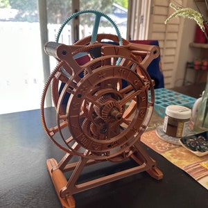 May include: A brown, intricate mechanical model with multiple gears and a zodiac wheel. The model has a complex design with a light blue gear at the top. It is displayed on a dark surface.