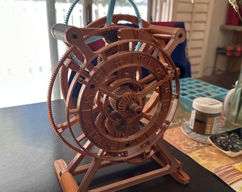 May include: A brown, intricate mechanical model with multiple gears and a zodiac wheel. The model has a complex design with a light blue gear at the top. It is displayed on a dark surface.