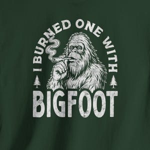 May include: Dark green t-shirt with a white graphic of Bigfoot smoking a cigar, accompanied by the text "I burned one with Bigfoot". The design includes two small pine trees on either side of the Bigfoot illustration.