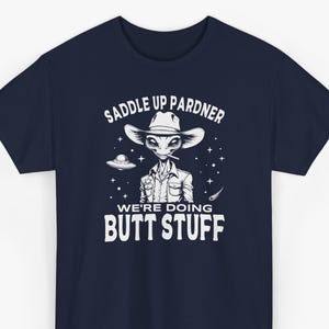 May include: A navy blue t-shirt featuring a white graphic of an alien wearing a cowboy hat and sheriff badge, with a flying saucer and stars. The text reads SADDLE UP PARDNER WE'RE DOING BUTT STUFF in bold white lettering centered on the chest.