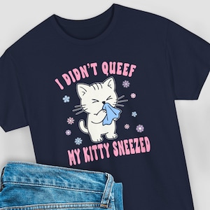 May include: Navy blue t-shirt with a graphic of a white cat holding a blue tissue, surrounded by flowers. Text reads "I DIDN'T QUEEF MY KITTY SNEEZED" in pink. The shirt is paired with blue jeans.