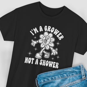 May include: Black t-shirt featuring a cartoon flower character with arms and legs. The flower has a smiling face and is wearing shoes. The text "I'M A GROWER NOT A SHOWER" is printed above and below the flower in white, retro-style lettering.