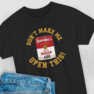 May include: Black t-shirt featuring a humorous graphic. The design includes a red and white can labeled "Grandpa's Condensed" with "Can of Whoop Ass" text. The words "Don't Make Me Open This!" are in a gold color.