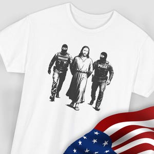 Puede incluir: Camiseta blanca con un gráfico en blanco y negro que representa una figura con túnica escoltada por dos figuras con equipo táctico. Las figuras están etiquetadas como "IMMIGRATION" y "POLICE". Una bandera estadounidense está en la esquina inferior derecha.