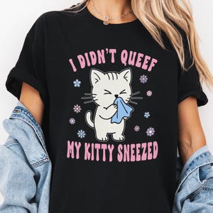 May include: A black t-shirt featuring a graphic of a white cat sneezing into a blue tissue. The text reads I DIDN'T QUEEF MY KITTY SNEEZED in pink letters. Small blue, purple, and white flower icons surround the cat on the front of the garment.