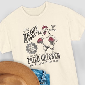 Può includere: T-shirt color crema con grafica in stile vintage. Il design include il testo "The Angry Rooster" e "World Famous Fried Chicken". È presente anche un gallo dei cartoni animati con guantoni da boxe. La maglietta ha anche il testo "You lookin' for a 2 piece?" e "Hot & Spicy!"
