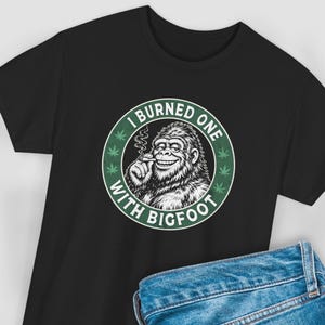 May include: Black t-shirt featuring a graphic of Bigfoot smoking a cigarette, with the text "I Burned One With Bigfoot" in a circular design. The design includes green cannabis leaves. The shirt is made of a soft material.