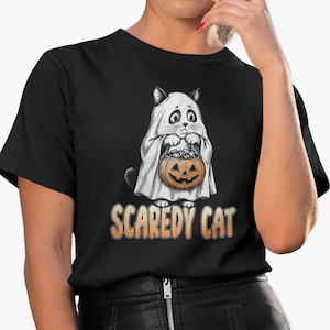 Pode incluir: Uma t-shirt preta de manga curta com um gráfico de um gato vestido de fantasma, segurando um balde de doces em forma de abóbora laranja. Abaixo da ilustração, o texto "SCAREDY CAT" está impresso numa fonte laranja com efeito degradê.
