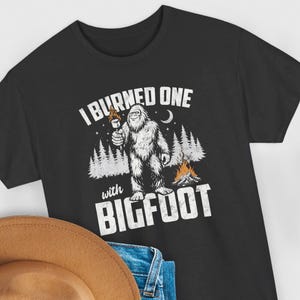 Puede incluir: Camiseta negra con un gráfico de Bigfoot. El diseño presenta un Bigfoot blanco sosteniendo una antorcha, una fogata y el texto "I burned one with Bigfoot."