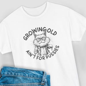 Könnte beinhalten: Weißes T-Shirt mit einer Schwarz-Weiß-Illustration einer Katze mit Brille und Gehstock. Der Text "Growing Old Ain't For Pussies" ist um die Katze herum gedruckt. Blaue Jeans sind unten zu sehen.