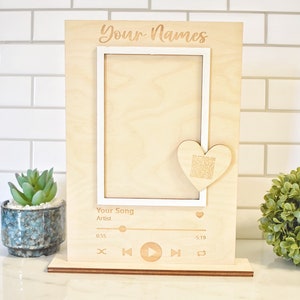 May include: A wooden photo frame with a white inner frame and a heart-shaped cutout. The frame has a music player design with a play button, rewind button, fast forward button, and a shuffle button. The text "Your Names" is engraved at the top of the frame. The text "Your Song" and "Artist" is engraved below the frame. The text "0:55" and "5:19" is engraved below the music player design.