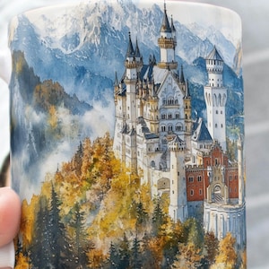 May include: White ceramic mug featuring a watercolor painting of a castle nestled in a mountain landscape. The artwork depicts a detailed castle with multiple towers, surrounded by autumn foliage and snow-capped mountains. The mug has a white handle.