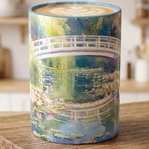 May include: A cylindrical ceramic mug featuring a colorful impressionist-style painting of a white arched bridge over a pond with water lilies. The mug is filled with a hot beverage topped with latte art, and steam rises from the top rim.
