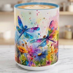 May include: A ceramic mug featuring a vibrant watercolor design of two dragonflies flying over a field of colorful wildflowers. The mug has a light blue interior rim and is filled with a hot beverage showing a latte art pattern on the surface.