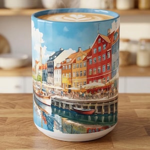 May include: A ceramic mug featuring a colorful illustration of a waterfront harbor with red, yellow, and blue buildings, a sailboat, and a small red boat. The mug is filled with a hot beverage topped with latte art, showing a light brown foam surface.