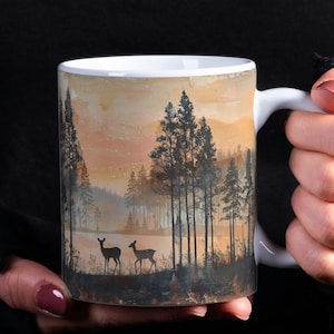 May include: White ceramic mug featuring a landscape scene with trees, a lake, and deer. The mug has a white handle and a light brown and orange background. The mug is held by a person wearing a black long-sleeved shirt.