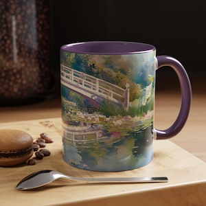 May include: A ceramic mug with a purple interior and handle, featuring a colorful watercolor-style print of a white arched bridge over a pond with water lilies and lush green foliage, reflecting the scene on the water surface below.