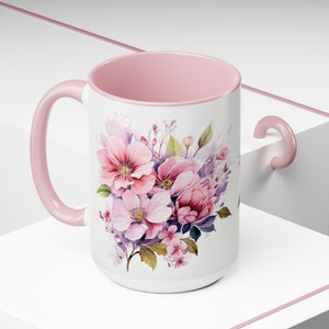 Puede incluir: Una taza de cer&aacute;mica rosa con un dise&ntilde;o floral blanco con flores rosas, moradas y verdes. La taza tiene un asa rosa.