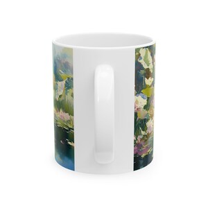 May include: A white ceramic mug featuring a wrap-around watercolor-style print of a pond scene. The artwork displays soft brushstrokes in shades of green, blue, pink, and white, depicting aquatic plants and reflections on the water surface.