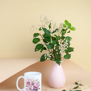 Puede incluir: Una taza de cer&aacute;mica blanca con un dise&ntilde;o floral y un jarr&oacute;n rosa con un ramo de flores blancas y hojas verdes sobre una superficie marr&oacute;n claro.