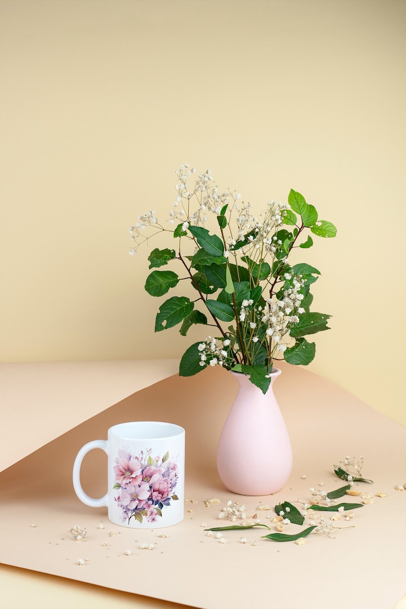 Puede incluir: Una taza de cer&aacute;mica blanca con un dise&ntilde;o floral y un jarr&oacute;n rosa con un ramo de flores blancas y hojas verdes sobre una superficie marr&oacute;n claro.