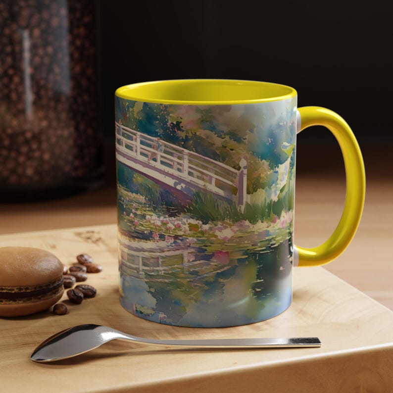 May include: A ceramic mug with a bright yellow interior and handle, featuring a colorful watercolor-style print of a white arched bridge over a pond with water lilies. The design depicts a serene garden scene with soft blue and green tones.