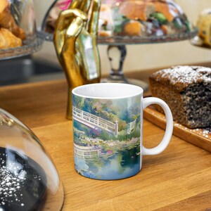 May include: A white ceramic mug featuring a colorful impressionist-style painting of a wooden footbridge over a pond with lush greenery and water lilies, with a handle on the right side and a smooth, glossy finish on the exterior surface.