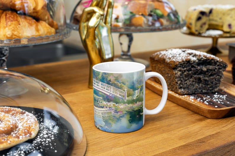 May include: A white ceramic mug featuring a colorful impressionist-style painting of a wooden footbridge over a pond with lush greenery and water lilies, with a handle on the right side and a smooth, glossy finish on the exterior surface.