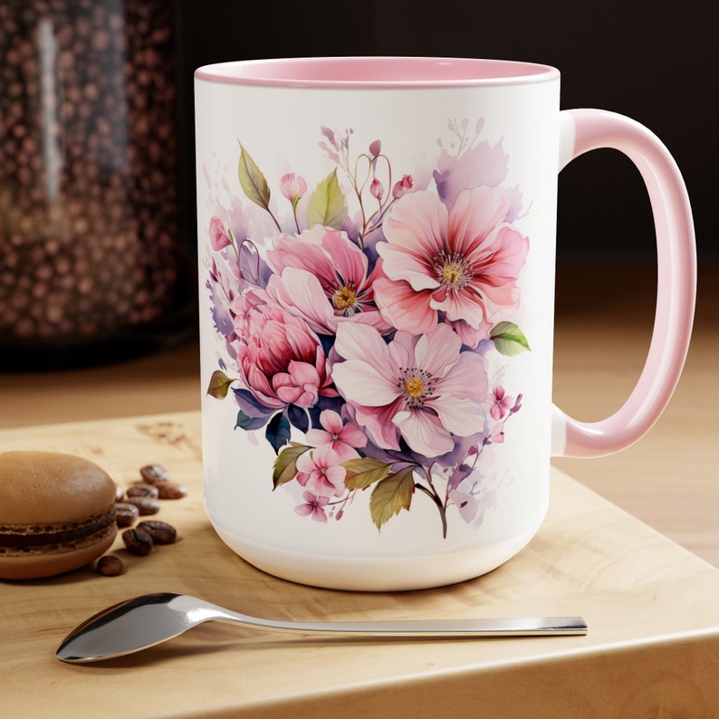 Puede incluir: Una taza de cer&aacute;mica blanca con un borde y un asa de color rosa presenta un dise&ntilde;o floral de acuarela con flores rosas, moradas y verdes.