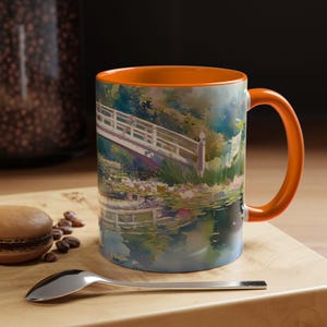 May include: A ceramic mug with a bright orange interior and handle, featuring a colorful watercolor-style print of a white arched bridge over a pond with water lilies and lush green foliage, capturing a serene garden scene in soft brushstrokes.