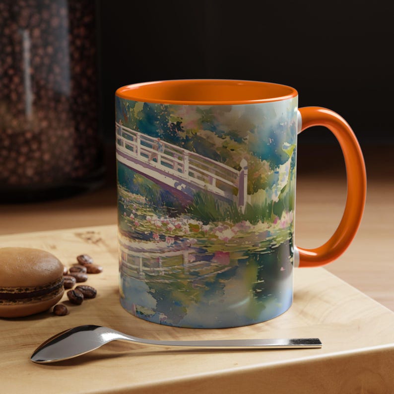 May include: A ceramic mug with a bright orange interior and handle, featuring a colorful watercolor-style print of a white arched bridge over a pond with water lilies and lush green foliage, capturing a serene garden scene in soft brushstrokes.