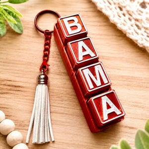 May include: A keychain featuring a red rectangular base with four raised square blocks, each displaying a white letter to spell BAMA. The keychain includes a red metal ring, a red chain, and a white faux leather tassel with a silver-toned cap.